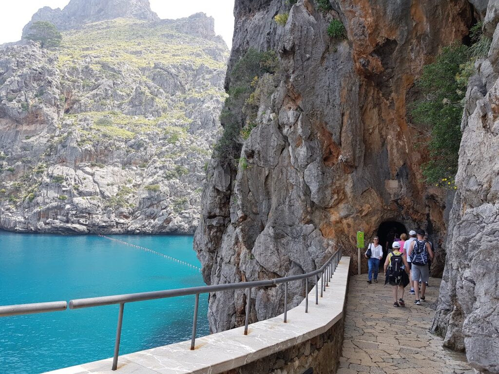 Mallorca Island Tour, Tourists walking along the coastal path through the Torrent de Pareis gorge in Sa Calobra, with turquoise Mediterranean water below.