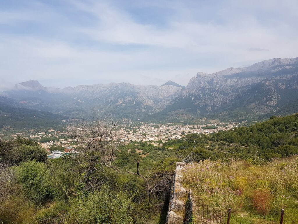 Mallorca Island Tour, Panoramic view of the town of Sóller, Mallorca, surrounded by lush green hills and the majestic Tramuntana mountain range in the background.
