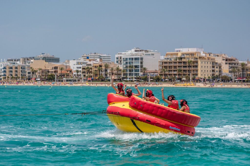 Participants having fun and waving while riding the thrilling Aqua Rocket Playa de Palma, with the lively beach promenade and buildings of Mallorca's Playa de Palma visible in the background.