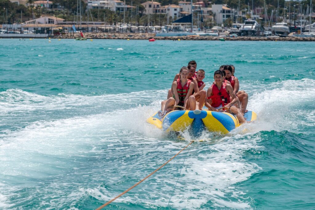 Group of young people riding a yellow and blue banana boat being towed across the turquoise waters at Playa de Palma, Mallorca. Boats, yachts, and waterfront buildings can be seen in the background.