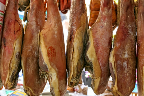 Several large, whole legs of cured ham (jamón) hanging in a traditional Spanish market. The hams, marked with seals of authenticity, are displayed in a row, showcasing their rich texture and aged appearance. The setup highlights the artisanal process of dry-curing meat, a hallmark of Spanish gastronomy, often found in local markets and delicatessens.