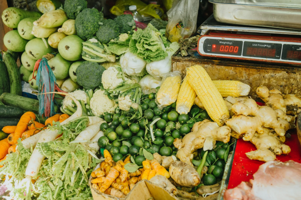 A vibrant market stall in weekly market Port de Soller , Mallorca, displaying an assortment of fresh vegetables and produce, including broccoli, cauliflower, lettuce, corn, carrots, cucumbers, radishes, limes, turmeric, ginger, and gourds. A weighing scale sits on the rustic wooden counter, adding to the authentic charm of this local farmers' market, where seasonal and organic ingredients are available for shoppers.