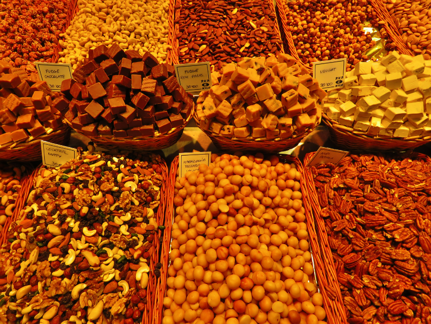 A warm-lit market stall featuring an assortment of dried fruits, nuts, and sweet treats. The display includes baskets filled with mixed nuts, almonds, pecans, nougat, chocolate fudge, and honey-roasted macadamia nuts. Handwritten price signs indicate the artisanal nature of the products. The rich golden hues and textures create an inviting and appetizing scene, reminiscent of a gourmet food market or specialty delicatessen.