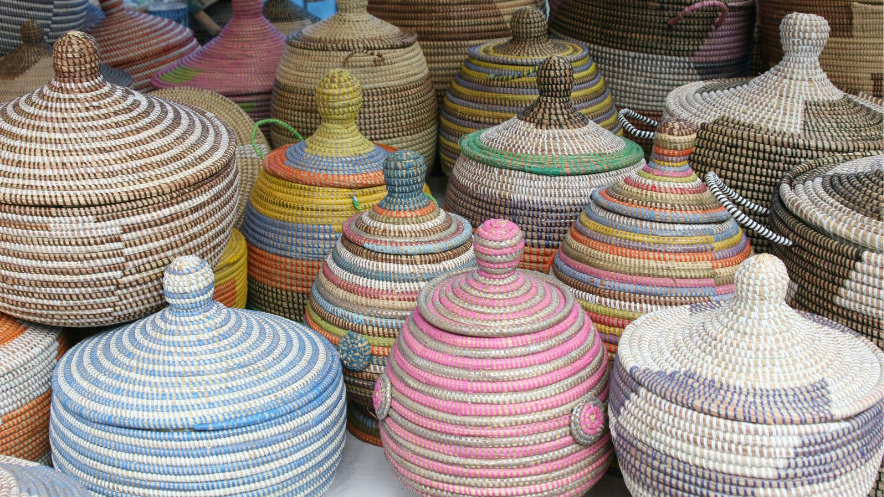 A collection of colorful, intricately woven baskets with lids displayed at a market stall. The baskets feature various patterns and color combinations, including blue, pink, yellow, and natural tones. Each basket has a unique design, with some featuring small handles and others having pointed lids. The image captures the craftsmanship of traditional handmade basketry, likely used for storage or decoration.