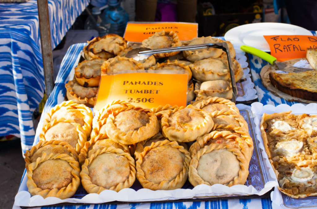 Golden-brown Mallorcan empanadas, known as panades, filled with traditional ingredients like Tumbet, meat, or fish, are carefully arranged at a market stall in Porreres, showcasing one of the island’s most beloved savory treats.