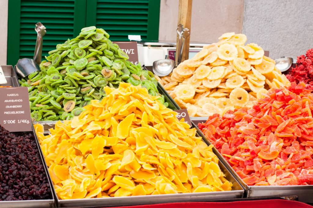 Brightly colored dried fruits, including green kiwi slices, golden mango, red papaya, and pineapple rings, are arranged in neat piles at a market stall in Porreres, Mallorca, creating a vibrant and enticing selection.