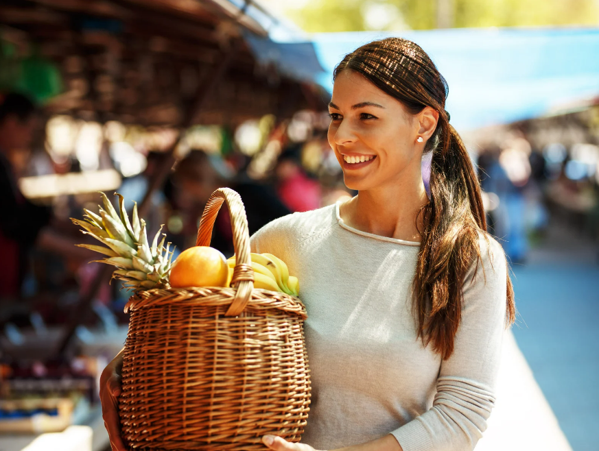A smiling woman with a basket full of fresh fruits at the Weekly Market Port de Sóller, surrounded by market stalls and a lively atmosphere.