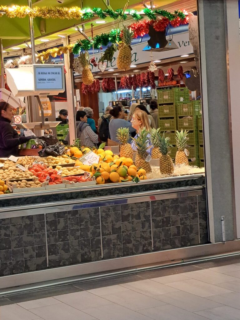 A fruit and vegetable stall inside Mercat de l'Olivar, decorated with pineapples and festive garlands, displaying fresh citrus and exotic fruits.
