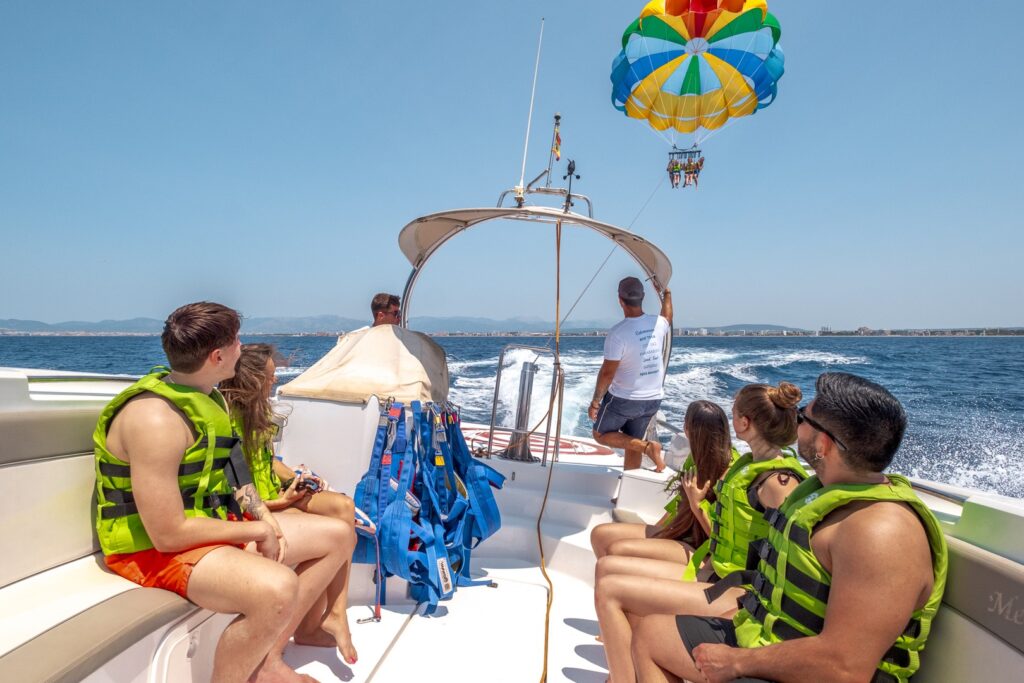 Group of people wearing life jackets sitting in a boat, watching others enjoying parasailing Playa de Palma, soaring high above the Mediterranean Sea off the coast of Mallorca.