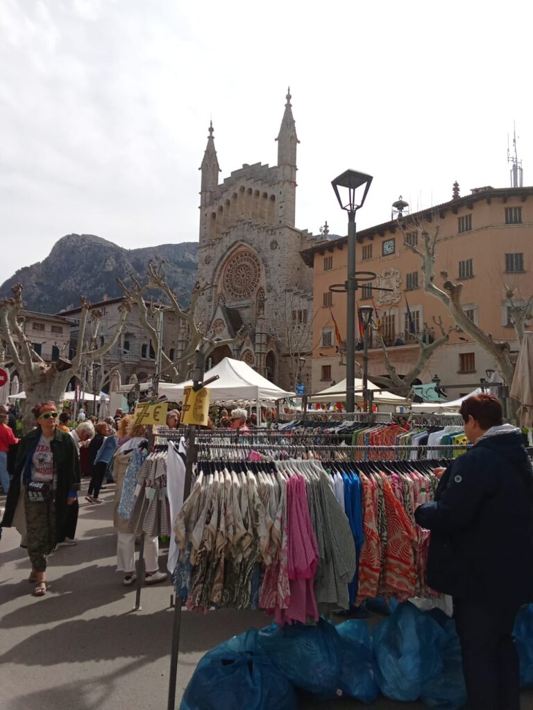 Bustling weekly market in Sóller with clothing racks and shoppers in front of the iconic Sant Bartomeu church, framed by the Tramuntana mountains.