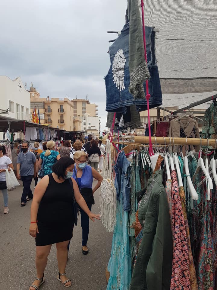 Visitors browsing colorful clothing stalls at the Weekly Market Arenal in Mallorca, with palm trees and market life in the background.