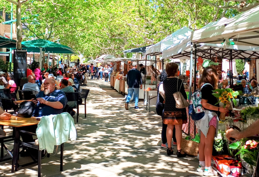 A lively scene at the Weekly Market in Esporles, Mallorca, with people browsing various stalls selling local goods, while others relax at outdoor cafés under lush green trees.
