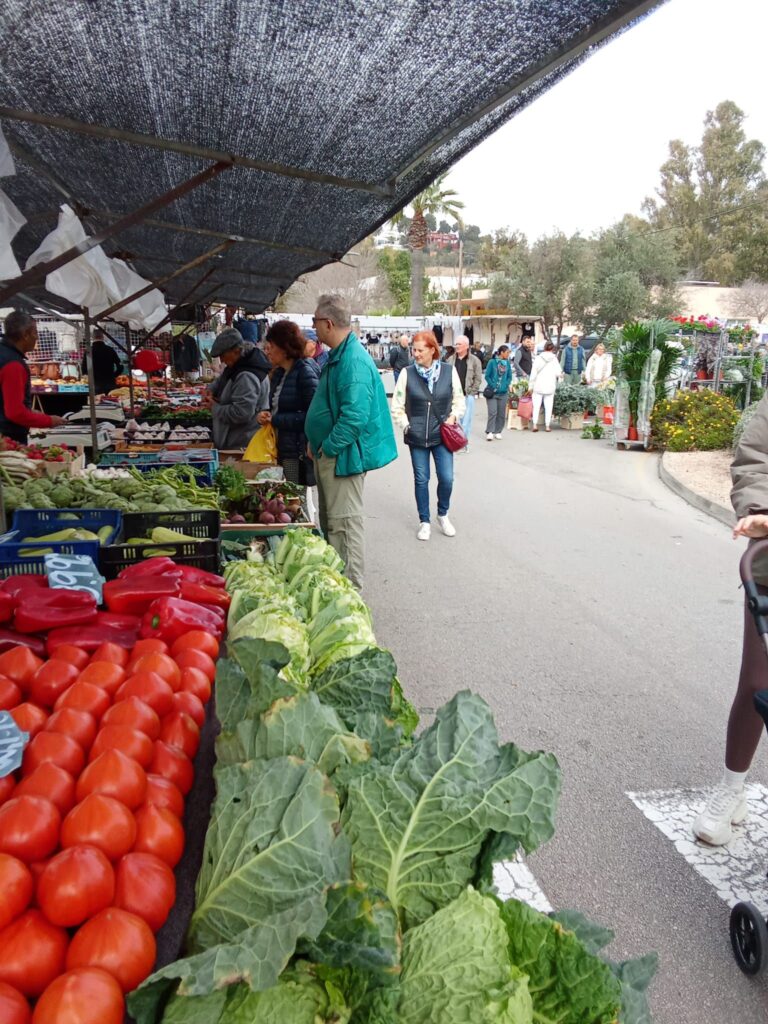 Visitors browsing fresh vegetables, including tomatoes, cabbage, and peppers, at the Weekly Market in Santa Ponsa, Mallorca, under a shaded canopy.