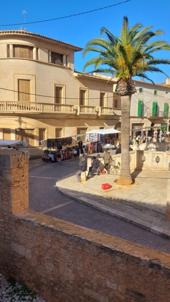 A scenic view of the Weekly Market Santanyí, with market stalls set against traditional Mallorcan architecture and a palm tree.