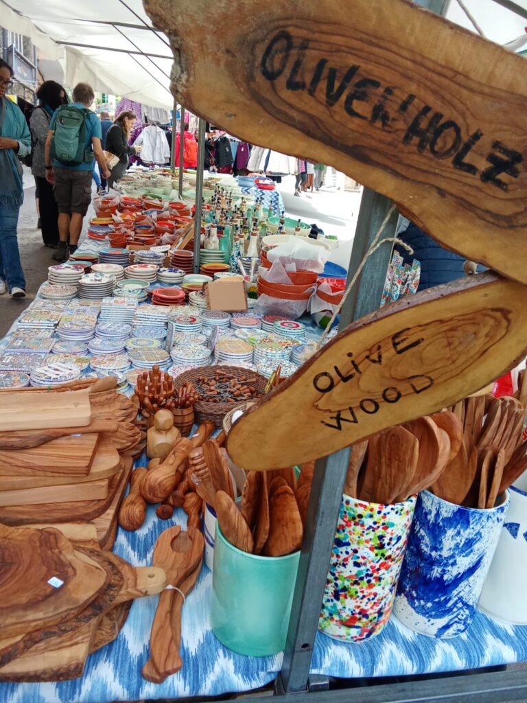 Market stall in Sóller offering handmade olive wood kitchen utensils, cutting boards, and colorful ceramic items, with a wooden sign reading “Olive Wood.
