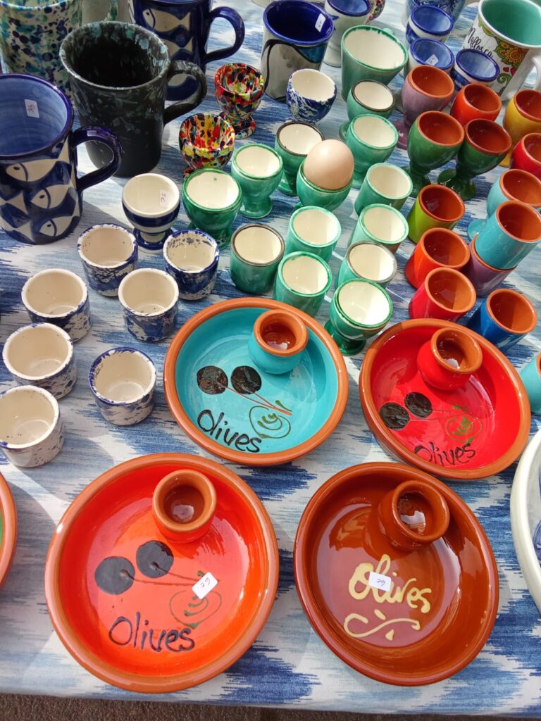 Brightly colored ceramic dishes labeled “Olives” alongside hand-painted cups and egg holders, artistically arranged on a blue-patterned tablecloth at the market.
