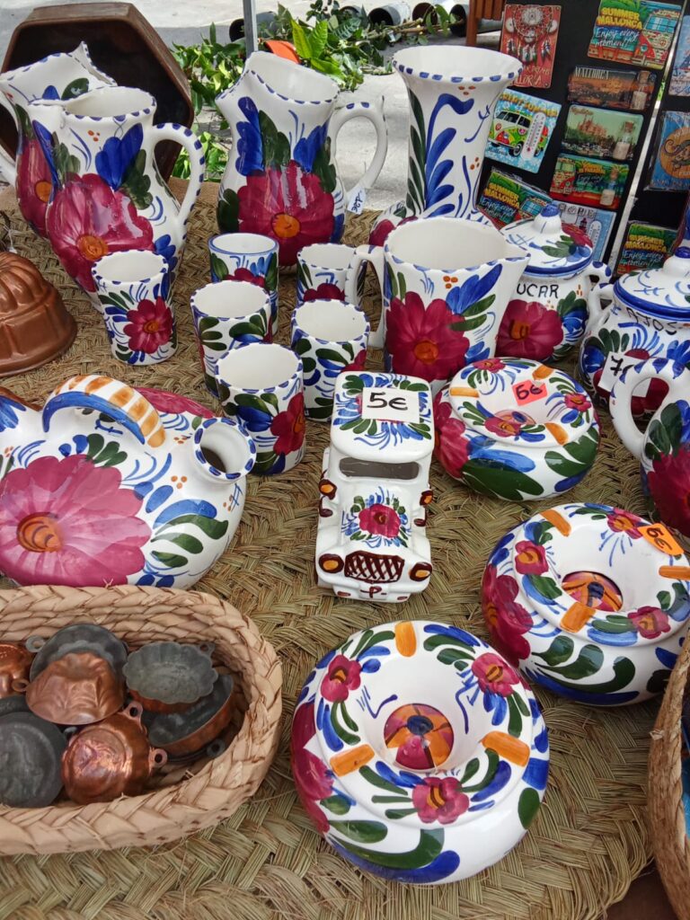 Vibrant hand-painted Mallorcan ceramics including jugs, cups, bowls, and decorative pieces with floral motifs, displayed on a woven mat at the Sóller market.