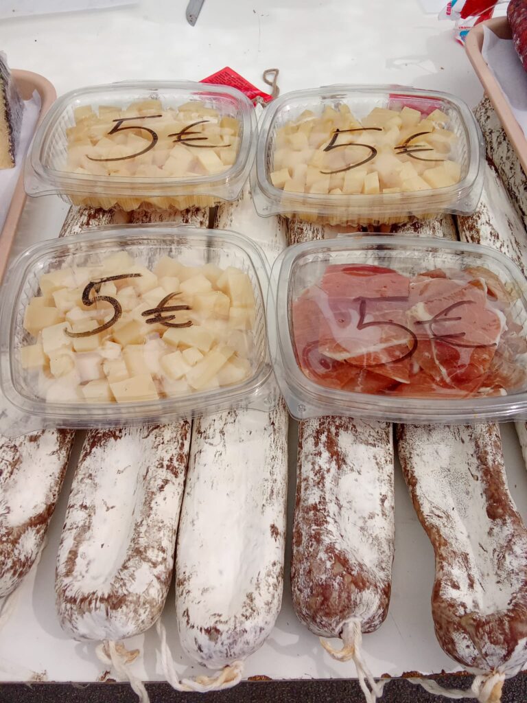 Packaged samples of local cheese and cured meats displayed over traditional Mallorcan sausage on a market table in Sóller, each container priced at €5.