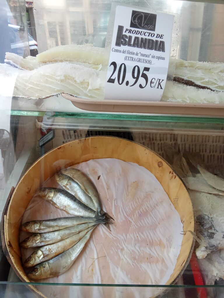 A glass display case showcasing salted cod fillets labeled as "Producto de Islandia," priced at 20.95€/kg, and a wooden tray with preserved fish at the Weekly Market Andratx in Mallorca.