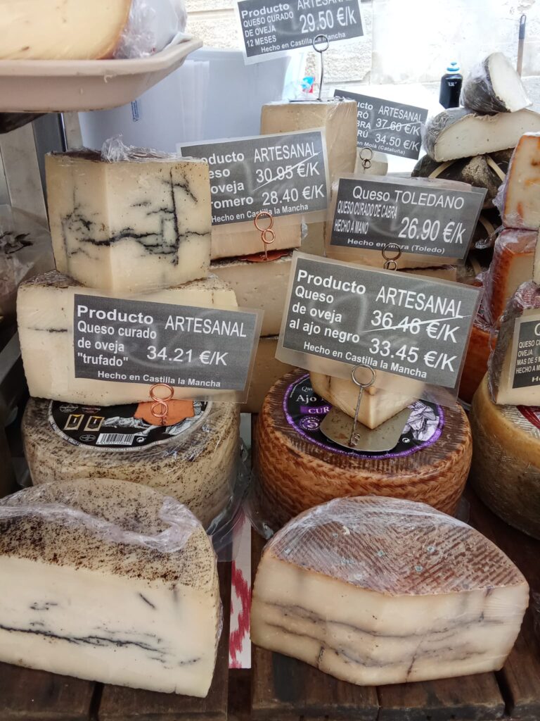 An assortment of artisanal cheeses at the Weekly Market Andratx in Mallorca, including truffle-infused sheep cheese, Manchego, and goat cheese, displayed with detailed pricing labels.