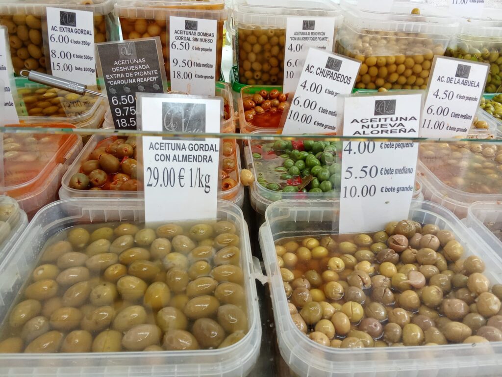 A selection of different types of olives at the Weekly Market Andratx in Mallorca, including spicy varieties and almond-stuffed olives, neatly arranged in transparent containers with price tags.