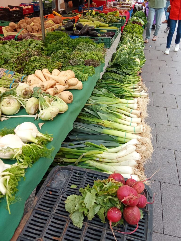 A vegetable stand at the Weekly Market Andratx in Mallorca displaying fresh produce, including leeks, radishes, parsnips, fennel, and leafy greens, arranged on a green tablecloth.