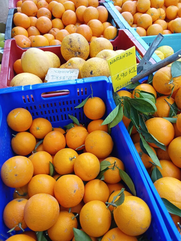 A vibrant fruit stall at the Weekly Market Andratx in Mallorca, featuring fresh oranges and grapefruits in colorful crates, with a sign indicating local Mallorcan oranges priced at 1.90€/kg.
