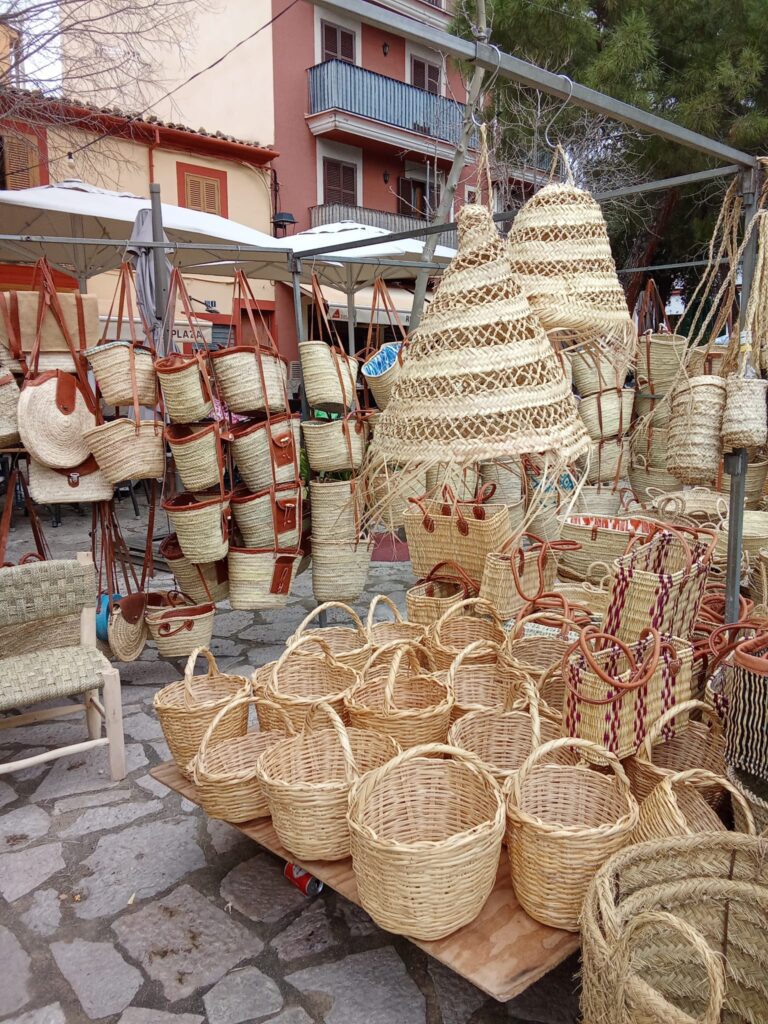 A collection of woven straw baskets, bags, and lampshades at the Weekly Market Andratx in Mallorca, crafted in traditional Mallorcan style and displayed in a charming outdoor setting.